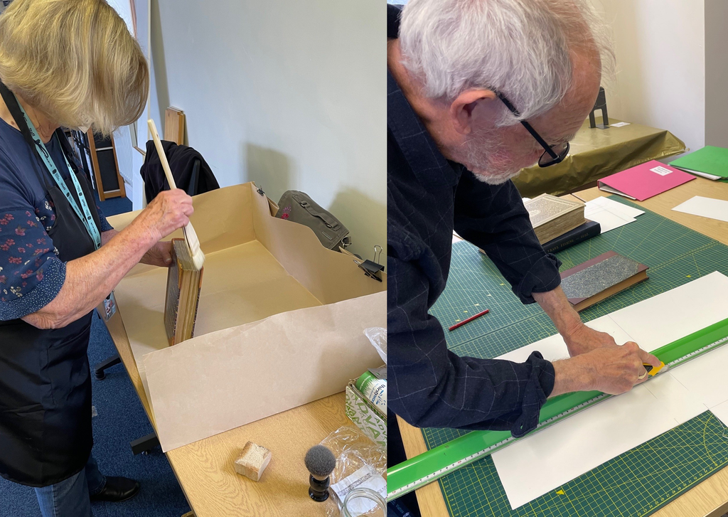 Kathy and Bernie doing book conservation work - Kathy is dusting the top of an old book with a white brush and Bernard is leaning over a cutting board, using a ruler and Stanley knife to cut a piece of cardboard lengthways.